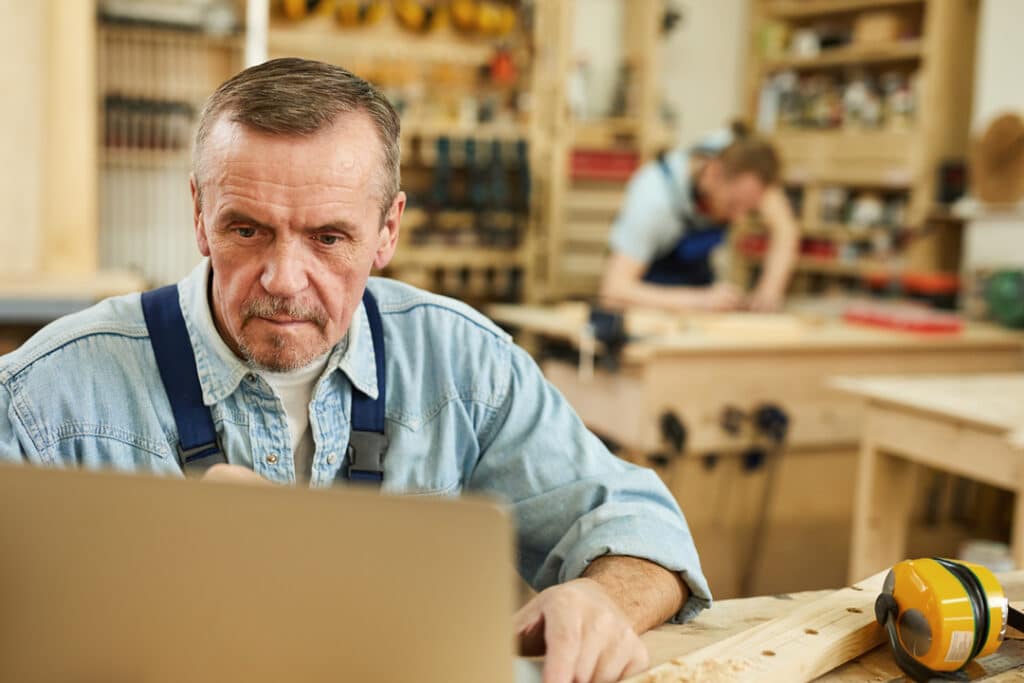 Portrait-of-senior-carpenter-using-laptop-while-working-in-joinery,-copy-space